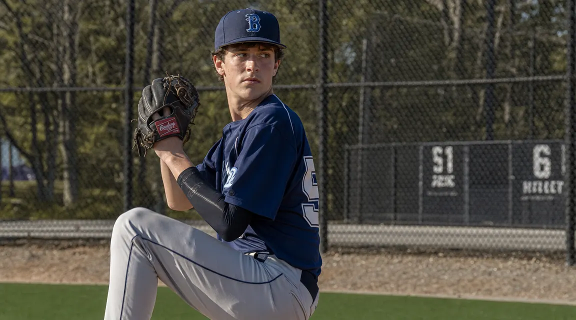 Teen baseball pitcher warming up in the bullpen with an elbow sleeve visible under his jersey on a sunny afternoon