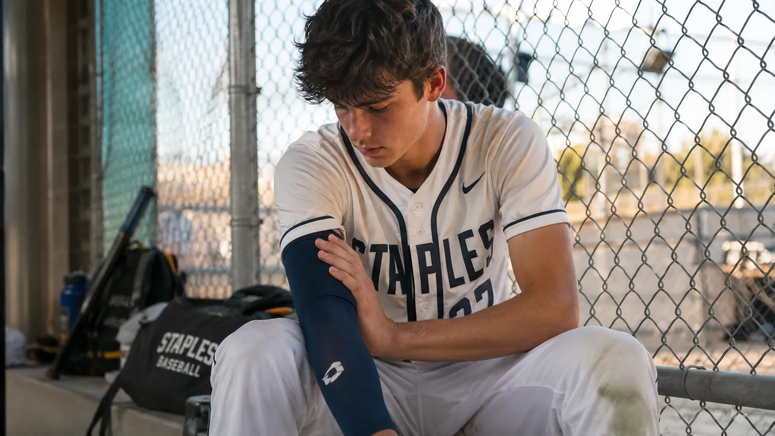Teen pitcher in dugout between innings with navy compression arm sleeve on his throwing arm, looking down at his elbow
