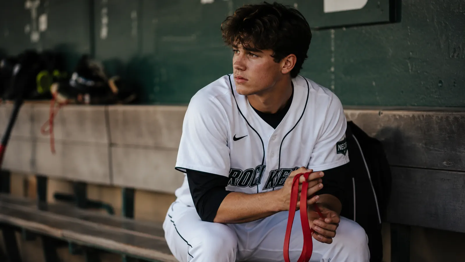 Teen pitcher holding Jaeger J-Bands resistance bands in a dugout, looking thoughtful before a start