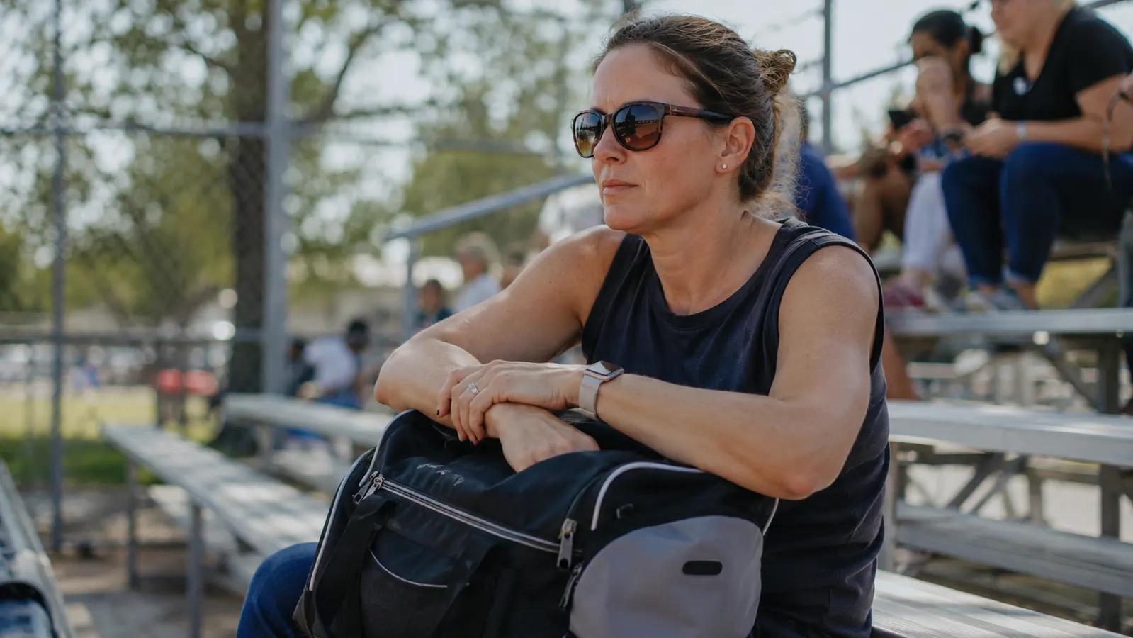 Mom sitting in bleachers watching a baseball game, looking attentive and calm, holding a gear bag on her lap
