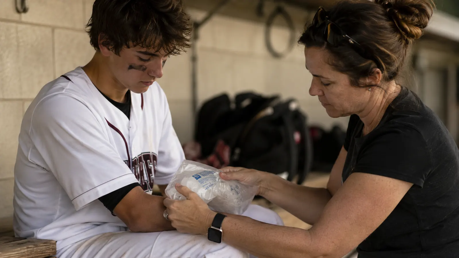 Mom applying ice wrap to son's elbow after a baseball game while he sits in the dugout