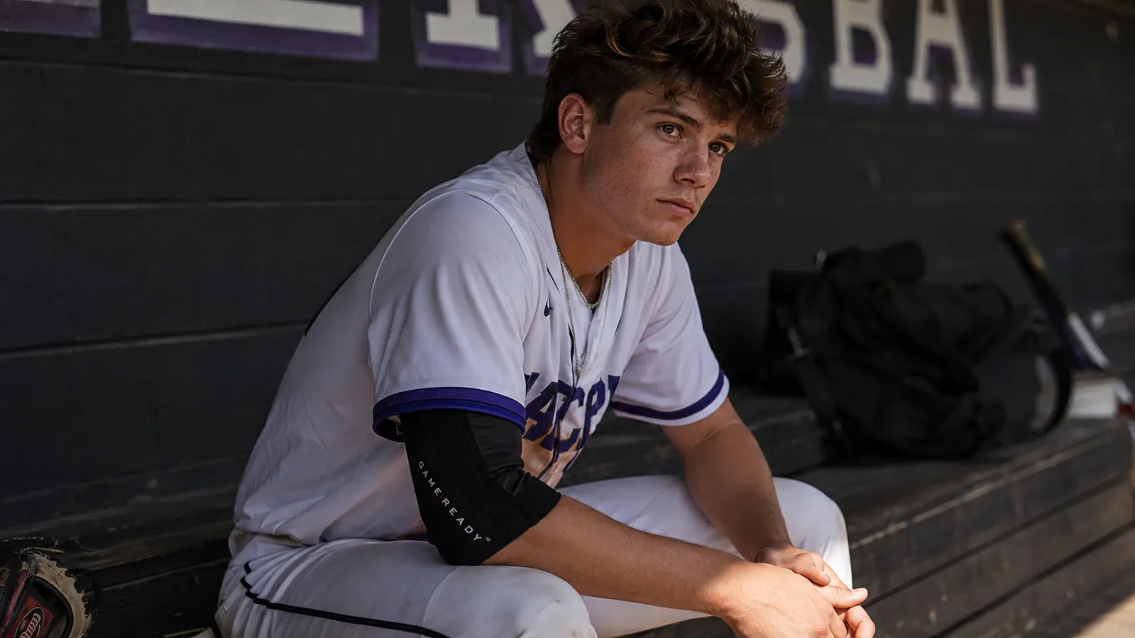 Teen pitcher sitting in dugout after a game, right elbow elevated, cold wrap applied, looking tired but calm